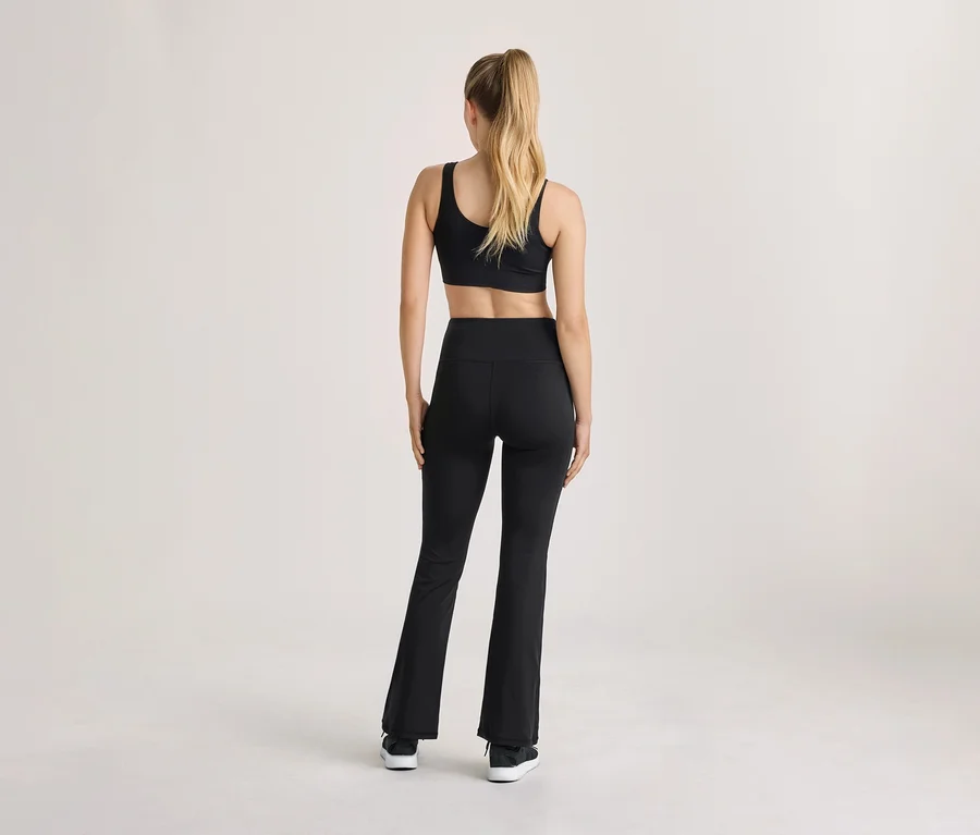 Femme de dos, portant une brassière de sport noire, un legging noir taille haute évasé et des baskets noires, les cheveux blonds attachés en queue de cheval.