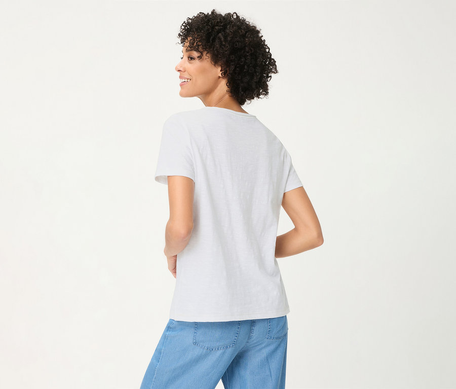Femme aux cheveux bouclés, vue de dos, portant un t-shirt Olsen blanc et un pantalon bleu clair, souriante.