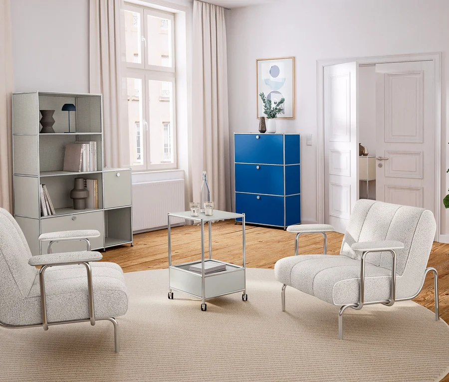 Salon lumineux avec parquet et murs blancs. À gauche, une étagère modulaire gris clair, au centre deux fauteuils clairs et une table d'appoint sur un tapis rond. À droite, une commode bleue et une plante.