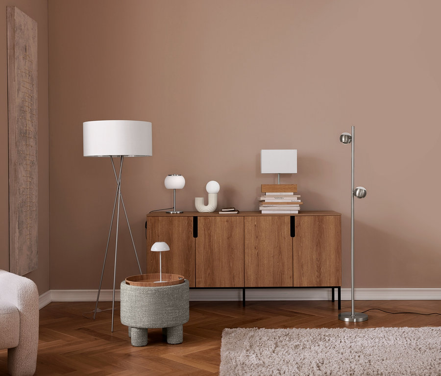 Un salon moderne avec des murs marron clair et un parquet en bois. Un fauteuil crème est à gauche. Un lampadaire trépied se tient à côté d'un pouf gris avec une petite lampe. Un buffet en bois contient deux lampes de table, un objet décoratif et des livres. Un lampadaire argenté à deux lumières sphériques est à droite, avec un tapis clair au sol.