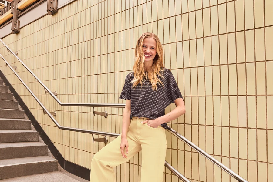 Une femme souriante aux longs cheveux blonds porte un haut à manches courtes à motifs sombres et un pantalon large jaune clair. Elle est appuyée contre une rampe à côté d'un mur carrelé et d'un escalier.