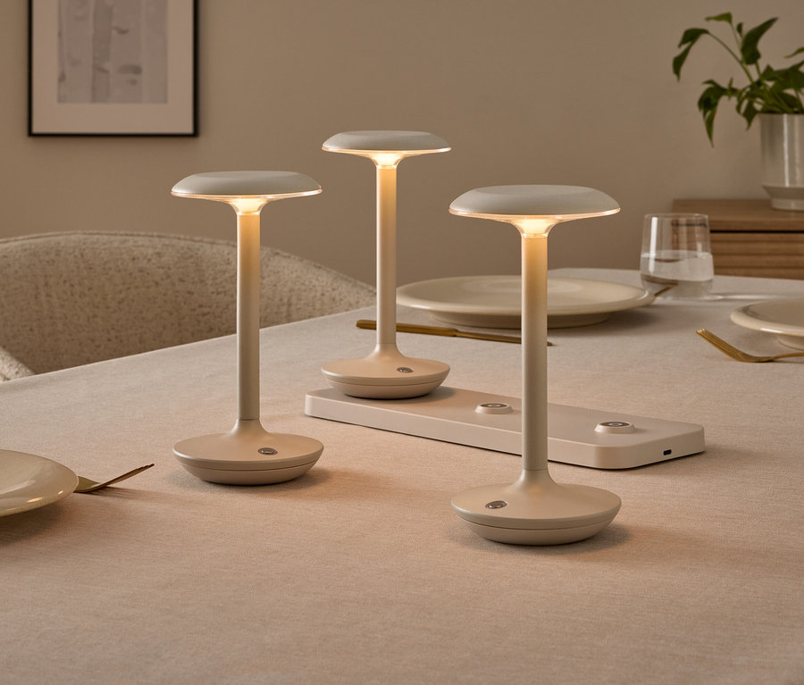 Trois lampes de table minimalistes de couleur crème sur une table à manger dressée, l'une d'elles sur une station de recharge. Les lampes émettent une lumière chaude.
