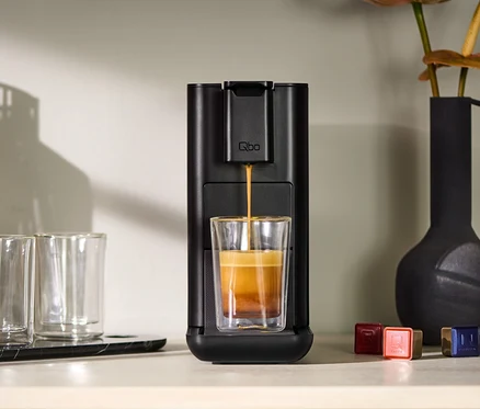 La machine à café Qbo ICONIC Dark Chrome remplit un verre de café. À côté se trouvent deux verres vides et un vase.
