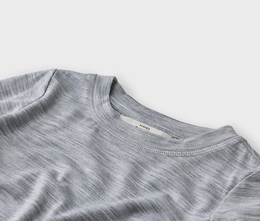 Gros plan d'un t-shirt gris chiné à col rond, montrant des coutures blanches visibles autour du col et une étiquette blanche avec le texte « TCHIBO ».