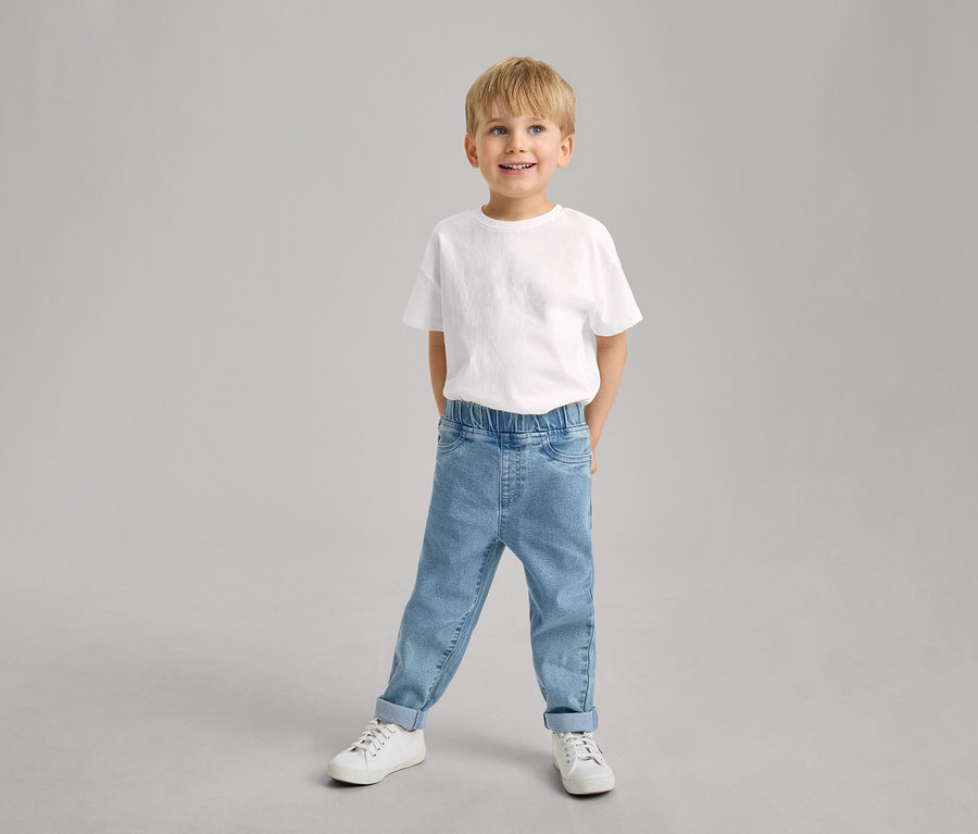 Un petit garçon souriant porte un T-shirt blanc, un pantalon à enfiler pour enfant – Fit « Luca » bleu clair avec les ourlets retroussés et des baskets blanches.
