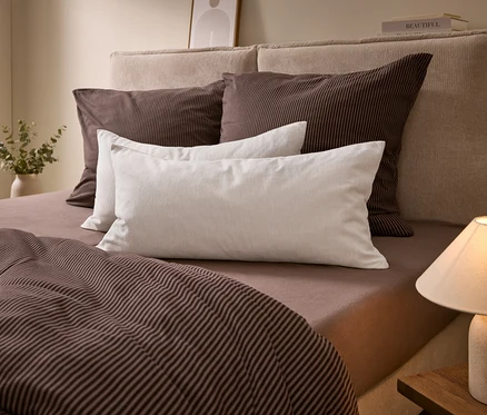 Un lit avec une tête de lit beige, une parure de lit rayée marron et des oreillers blancs. Une lampe est sur une table de chevet, un vase avec de la verdure à gauche.