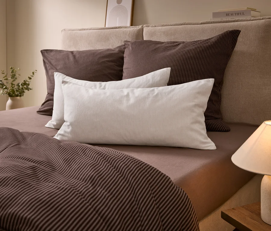 Un lit avec une tête de lit beige, une parure de lit rayée marron et des oreillers blancs. Une lampe est sur une table de chevet, un vase avec de la verdure à gauche.