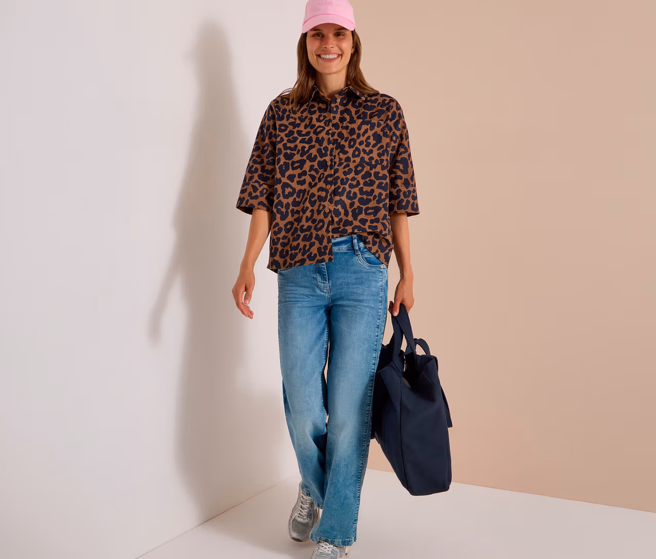 Femme souriante portant une casquette rose, un jean bleu clair, des baskets argentées et un chemisier CECIL à imprimé léopard marron et noir, tenant un sac fourre-tout bleu foncé.