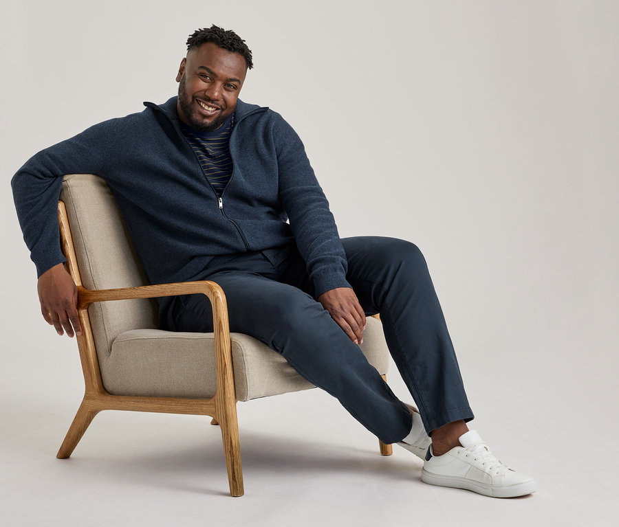 Homme souriant assis dans un fauteuil, portant une veste zippée bleu marine, un t-shirt rayé, un pantalon chino bleu marine et des baskets blanches.