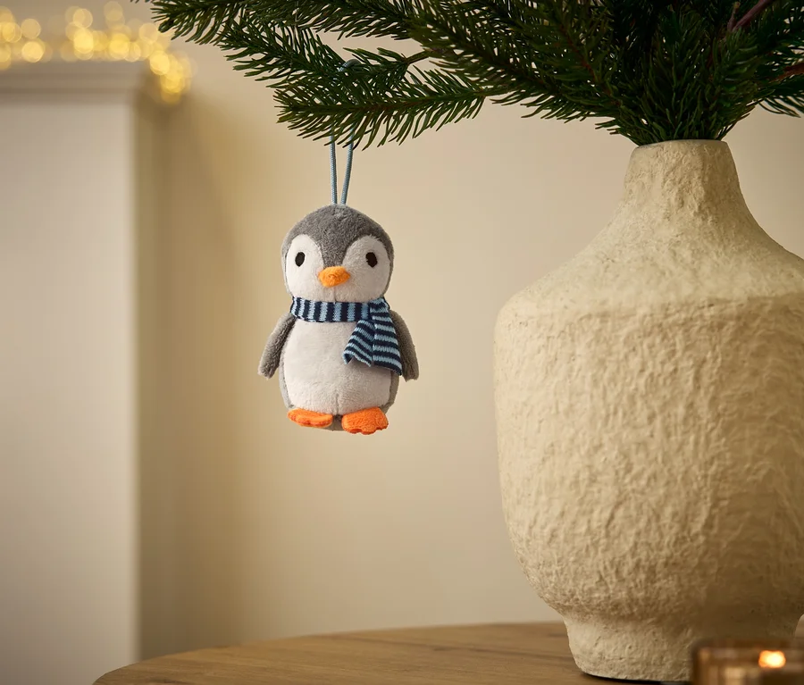 Un ornement pingouin en peluche gris et blanc avec un bec et des pieds orange, portant une écharpe rayée bleue et blanche, est suspendu à une branche verte. Un vase texturé de couleur claire avec des branches se trouve à côté, avec des lumières floues en arrière-plan.