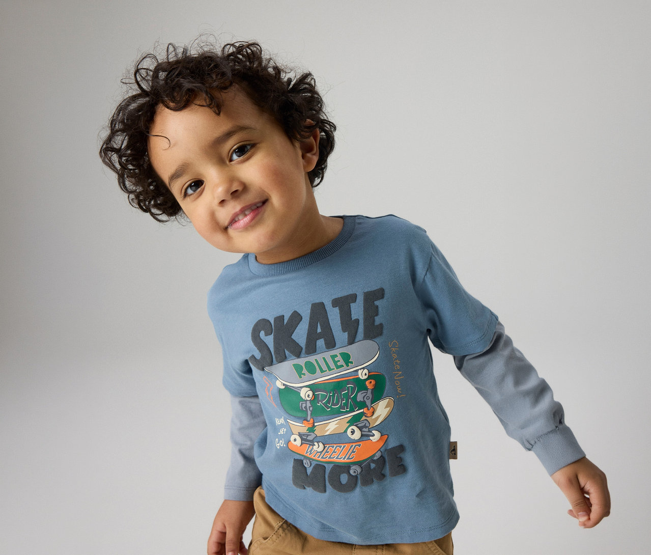 Jeune garçon souriant aux cheveux bouclés foncés, portant un t-shirt bleu à manches longues avec un motif "SKATE" et des skateboards empilés, et un pantalon marron, regardant la caméra.