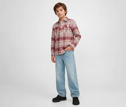 Jeune garçon debout, portant une chemise à carreaux rouge, blanche et beige à manches longues, un jean ample bleu clair et des chaussures noires. Il a une main dans sa poche.