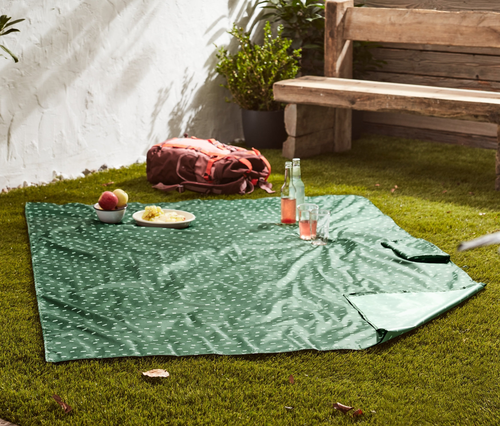 Une couverture de pique-nique 2 en 1 verte avec un motif blanc est étendue sur l'herbe. Des assiettes avec de la nourriture et des boissons sont posées sur la couverture.