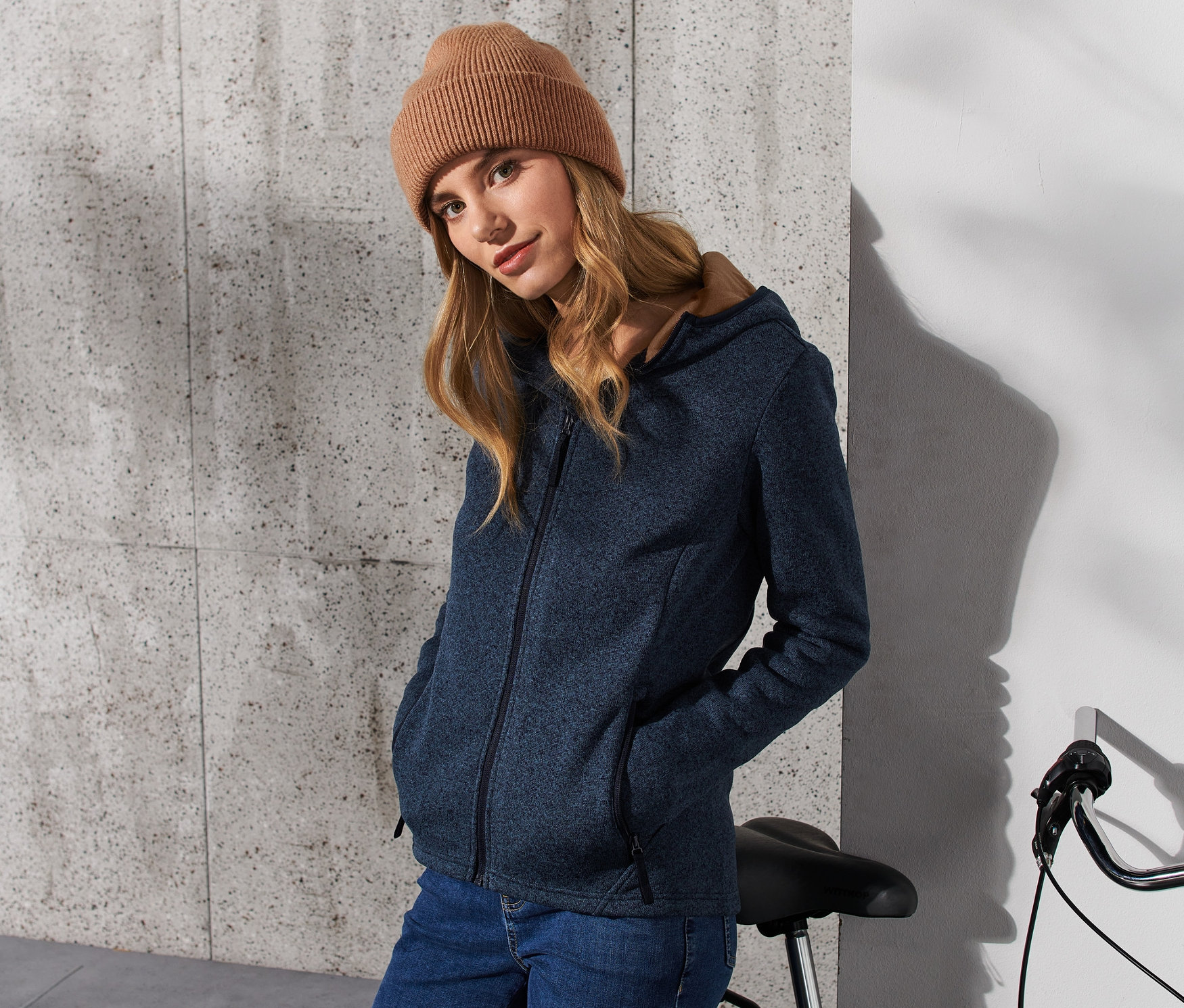 Une femme porte une veste en maille polaire bleu foncé et un bonnet marron, elle se tient à côté d'un vélo.