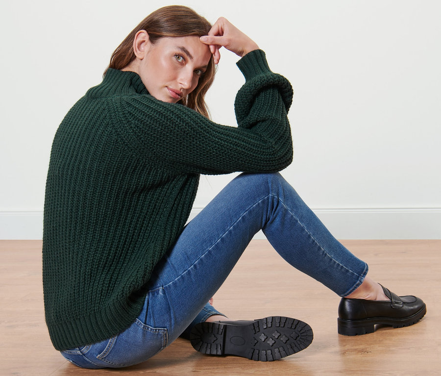 Une femme est assise par terre et porte un pull en maille vert foncé à col montant, un jean et des mocassins noirs.
