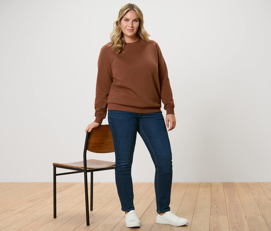 Une femme se tient à côté d'une chaise et porte un sweat-shirt marron et un jean slim – Coupe « Emma », bleu foncé.