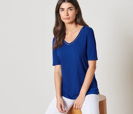 Une femme est assise sur un tabouret et porte un haut bleu cobalt et un pantalon blanc.