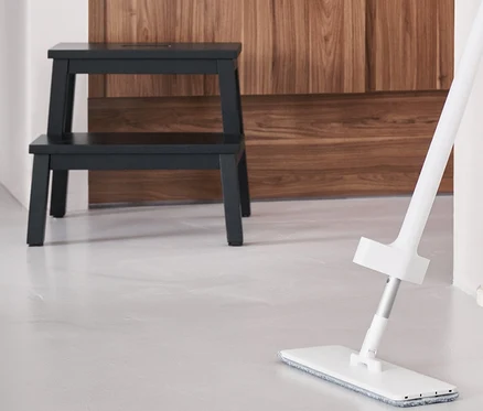 Un balai de sol avec spray est posé sur le sol, à côté d'un seau pliable. À l'arrière-plan se trouve un tabouret à 2 marches.