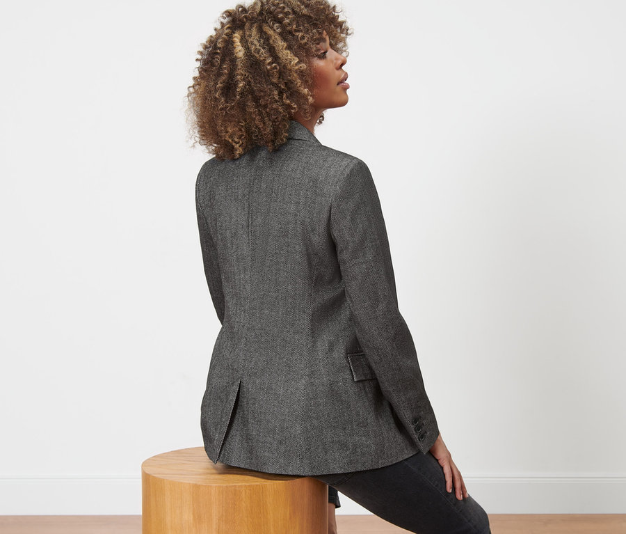 Une femme est assise sur un tabouret en bois et porte un blazer tissé aspect chevrons.