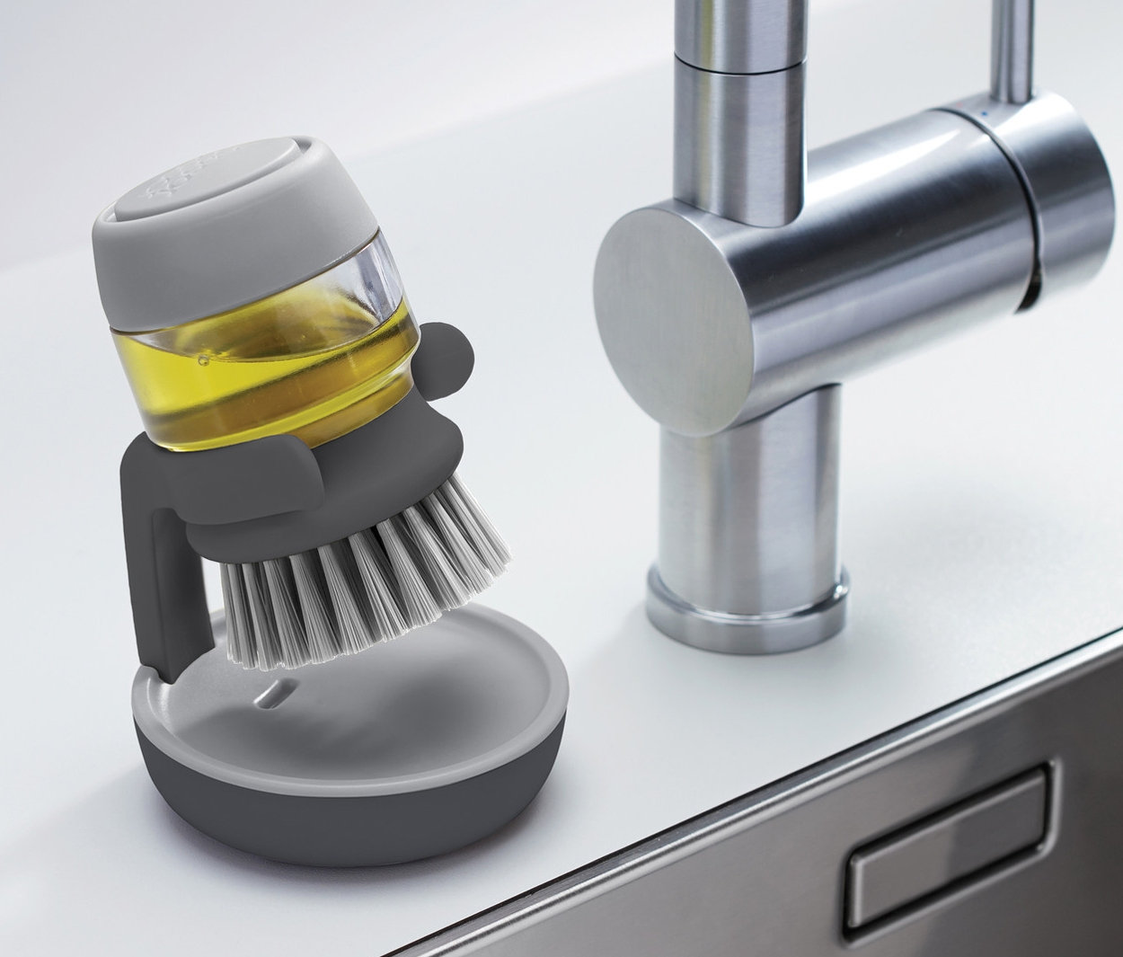 Une brosse à vaisselle grise Joseph Joseph avec distributeur de savon se trouve à côté d'un évier en acier inoxydable.