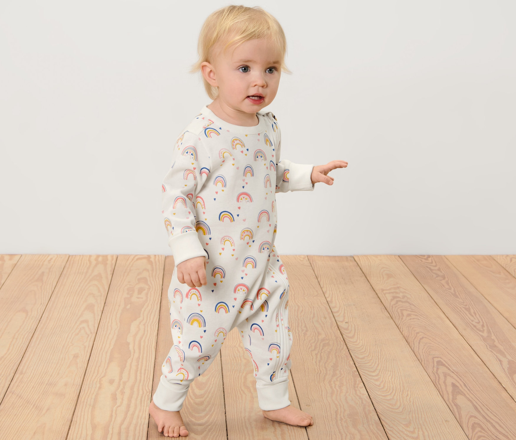 Un bébé en pyjama à imprimé arc-en-ciel se tient sur un plancher en bois. 2 pyjamas pour bébé, arc-en-ciel