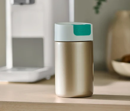 Un gobelet doré Kambukka To-Go est posé sur une table à côté d'une machine à café.