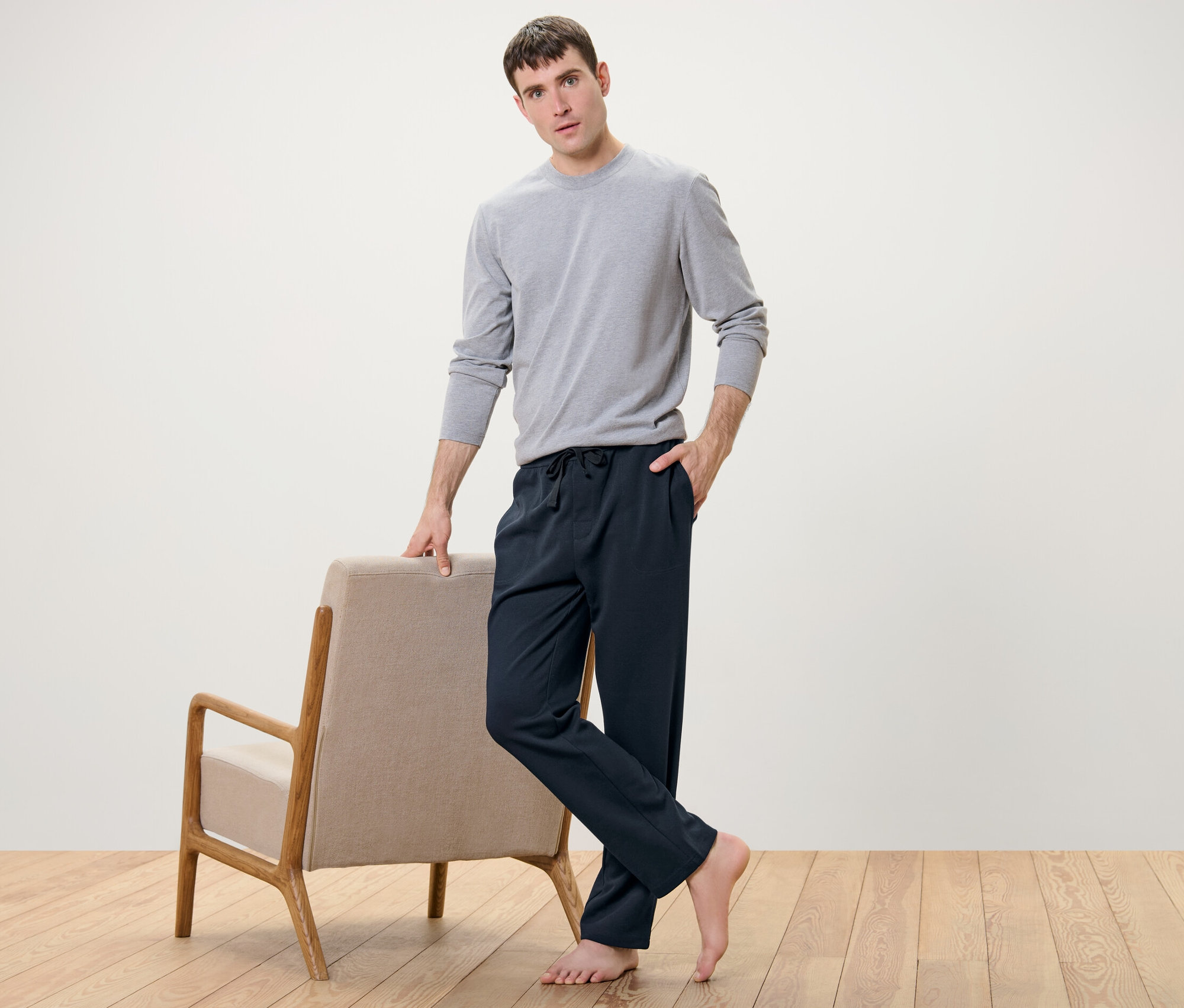 Un homme pose à côté d'un fauteuil, vêtu d'un pantalon d’intérieur et d'un t-shirt gris.