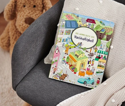 Sur un fauteuil gris reposent un plaid en tricot beige et le livre « Das Wimmelbuch der Nachhaltigkeit ». Un ours en peluche est visible en arrière-plan.