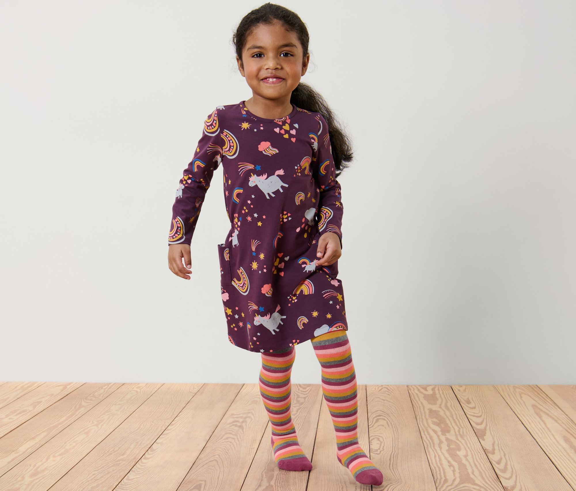 Une fillette souriante pose dans une robe licorne violette et des collants thermiques aubergine à rayures multicolores.