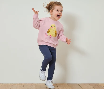 Une fille saute sur un plancher en bois, portant un sweat-shirt pour enfant avec intérieur gratté et des leggings.