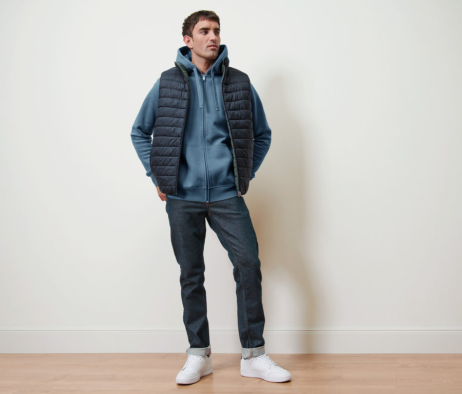 Un homme pose vêtu d'une veste à capuche bleu fumée, d'un gilet réversible et d'un jean.