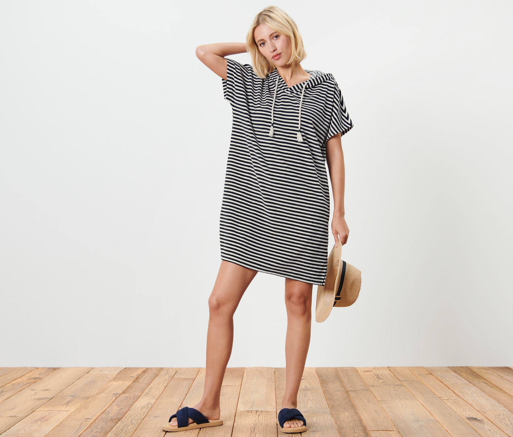 Une femme blonde pose en robe de plage en tissu éponge rayée bleu et blanc avec capuche, tenant un chapeau de paille et portant des mules bleues.