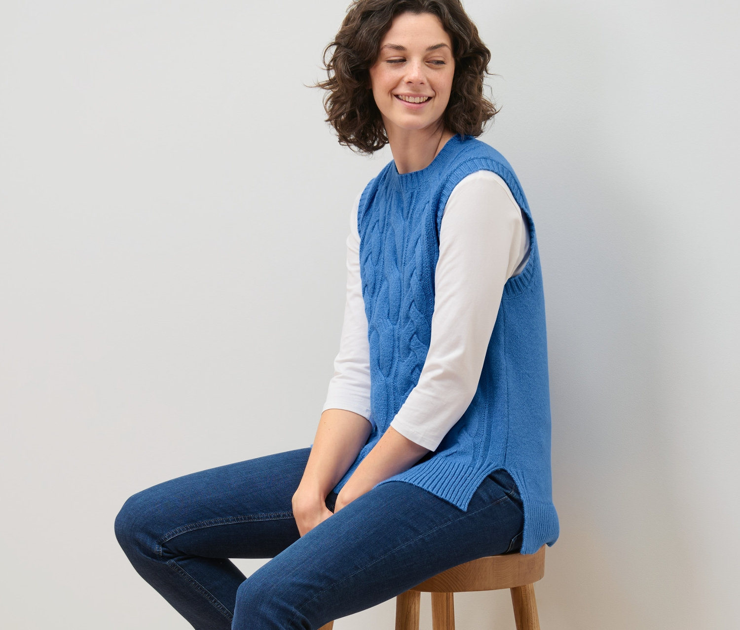 Femme assise sur un tabouret portant un pull sans manches en maille à motif torsadé.
