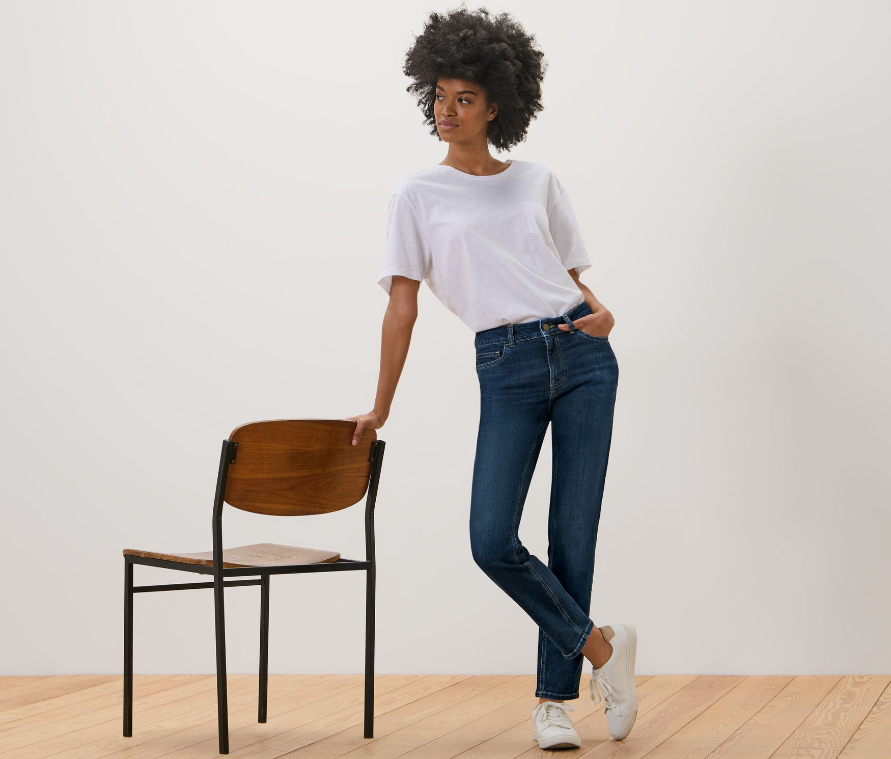 Une femme pose à côté d'une chaise et porte un jean slim – Coupe « Emma », bleu foncé.