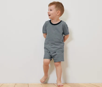 Un garçon en pyjama court 2
Pyjamas pour enfant, court, se tient pieds nus sur un plancher en bois à côté d'un ours en peluche.
