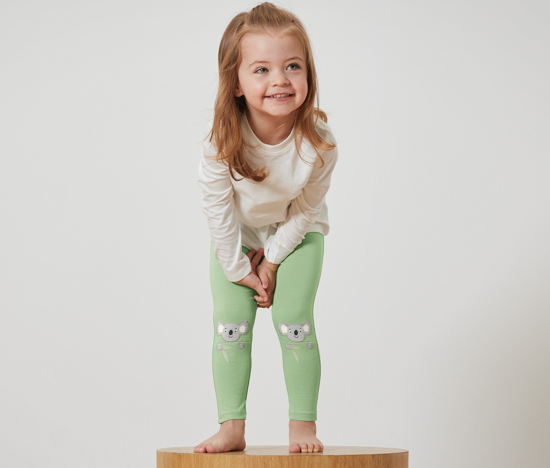 Une fille se tient sur un socle en bois et porte des leggings verts avec des motifs de koala et un t-shirt blanc.