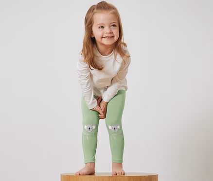 Une fille se tient sur un socle en bois et porte des leggings verts avec des motifs de koala et un t-shirt blanc.