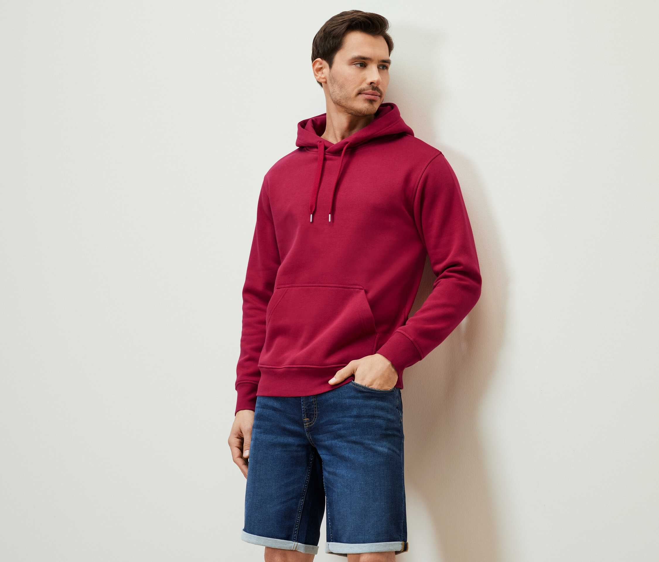 Un homme pose en sweat à capuche rouge et short en jean.
