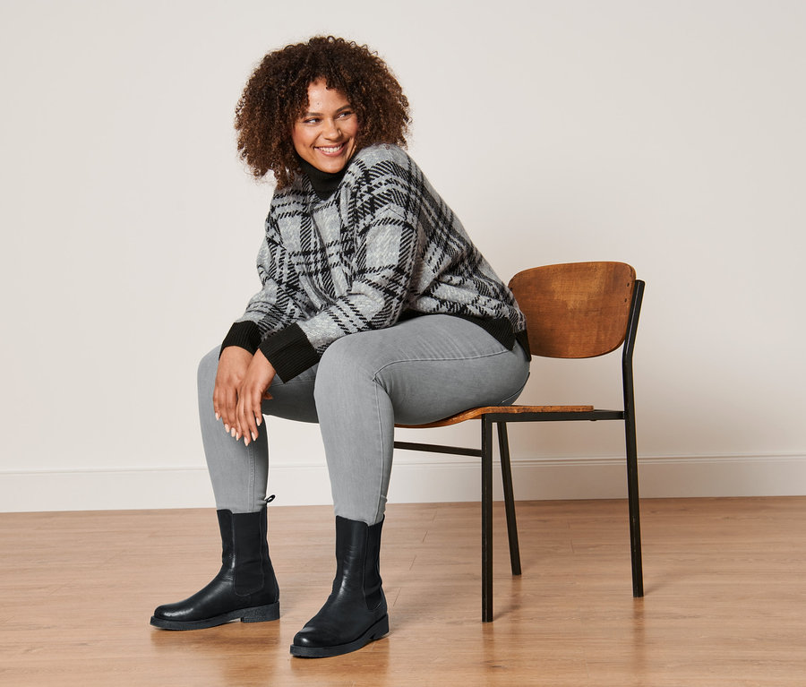 Une femme est assise sur une chaise et porte un pull en maille à col roulé, un tregging gris chiné et des bottines en cuir.