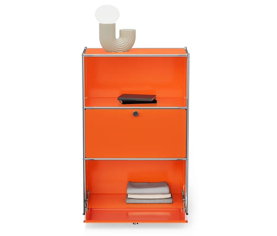 Meuble de rangement orange à trois compartiments avec cadre argenté. Une lampe beige clair et un livre noir sont posés sur le compartiment supérieur. Le compartiment inférieur est ouvert et contient des tissus pliés.