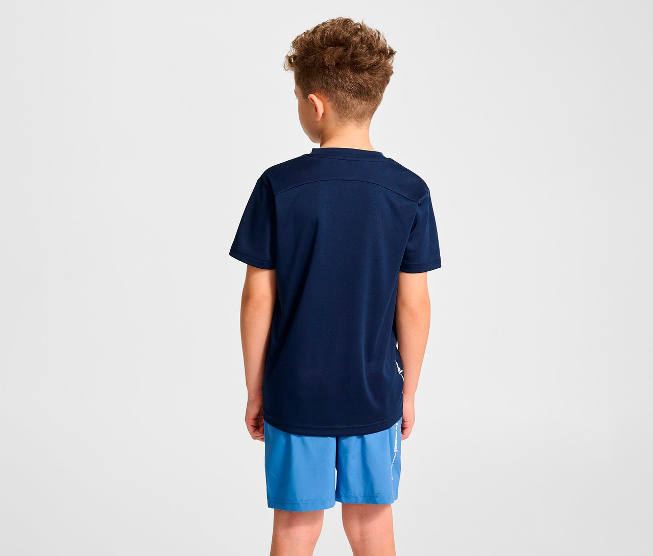 Un garçon, vu de dos, porte un t-shirt d’entraînement HUMMEL HMLJR pulse bleu foncé et un short bleu.