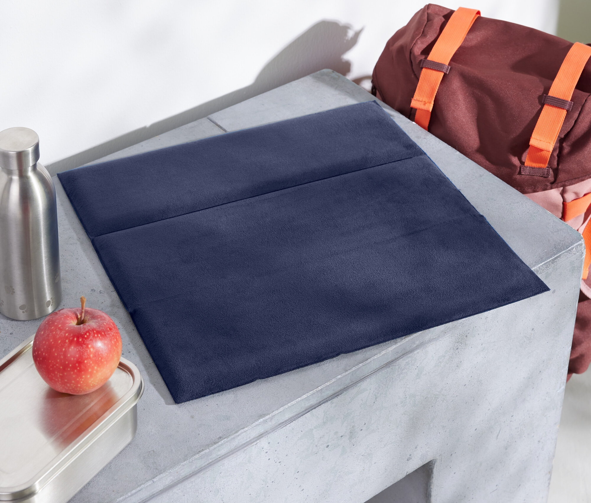 Sur une table en béton, on trouve un dessous de plat en tissu bleu, une pomme, une boîte en inox et une bouteille. À l'arrière-plan, il y a un sac à dos marron avec des bretelles orange.