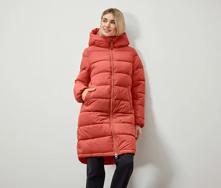 Une femme porte un long manteau matelassé à capuche et a les mains dans sa poche.