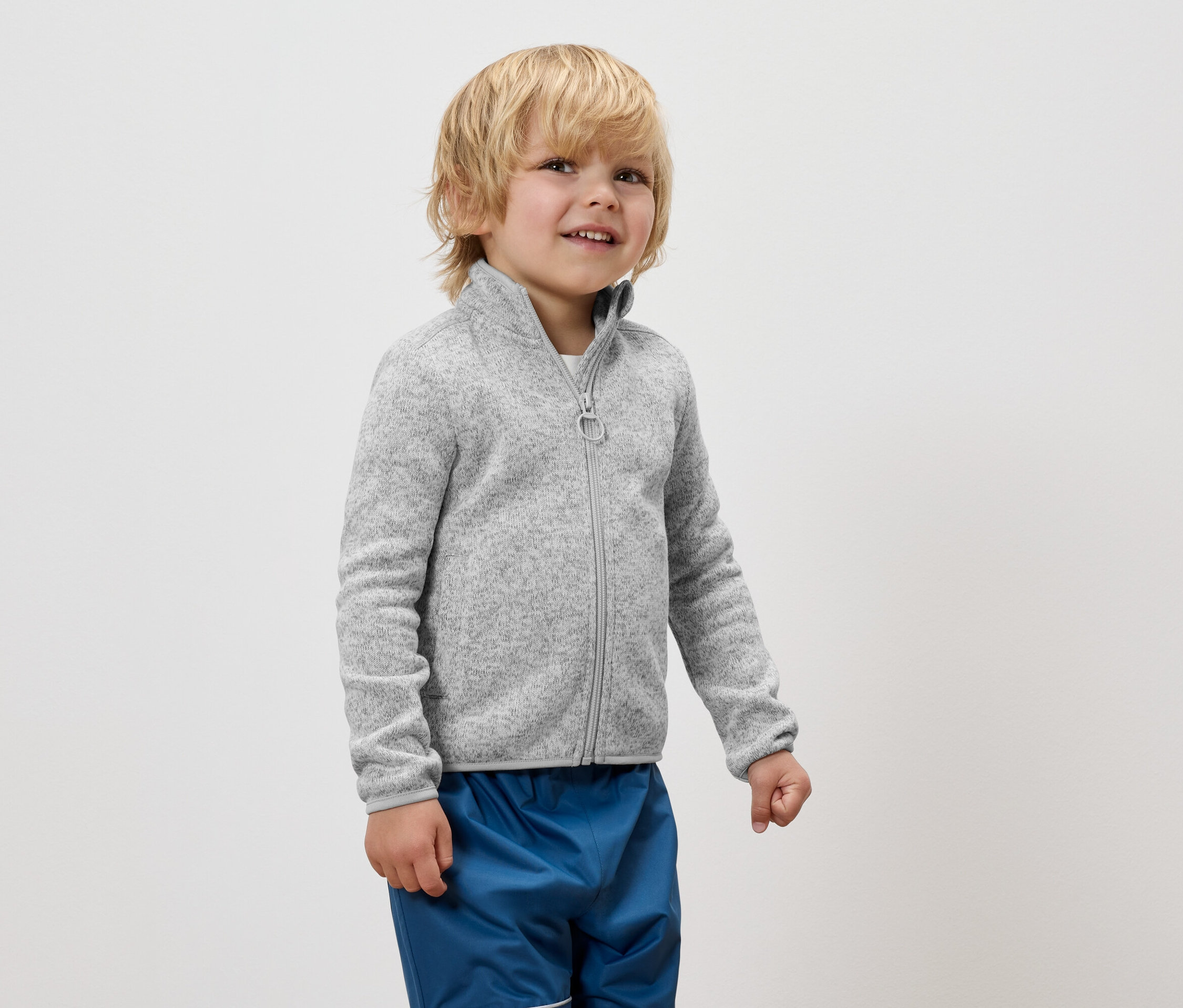 Un garçon portant une veste en polaire grise zippée et un pantalon bleu se tient sur un fond blanc.