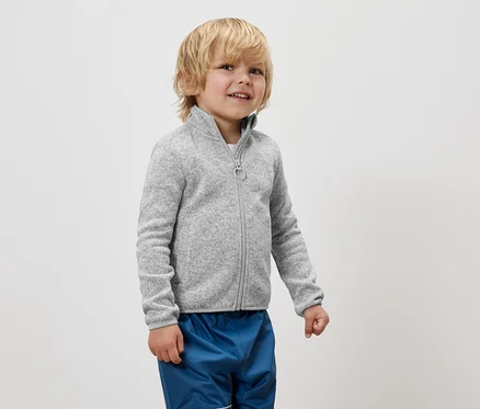Un garçon portant une veste en polaire grise zippée et un pantalon bleu se tient sur un fond blanc.