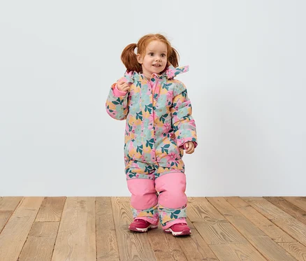 Une fille rousse en combinaison fleurie et chaussures roses se tient sur un plancher en bois devant un mur blanc.