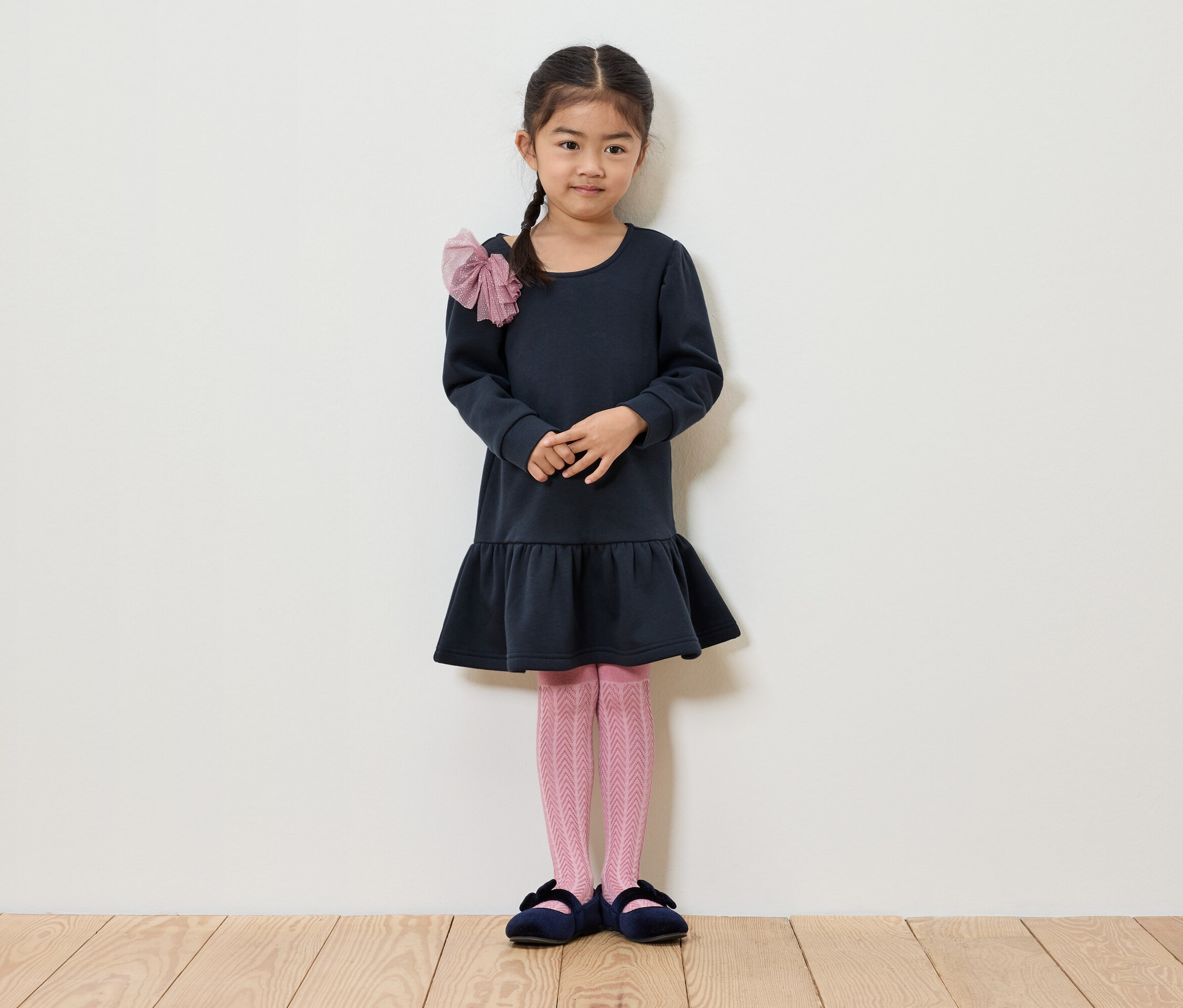 Une fille avec une tresse se tient devant un mur blanc. Elle porte une robe bleu foncé, des collants roses et des chaussures bleues.