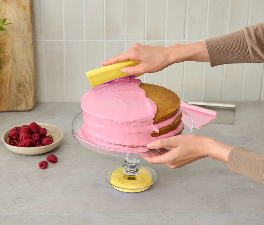 Une personne décore un gâteau rose sur un présentoir en verre avec un outil jaune.