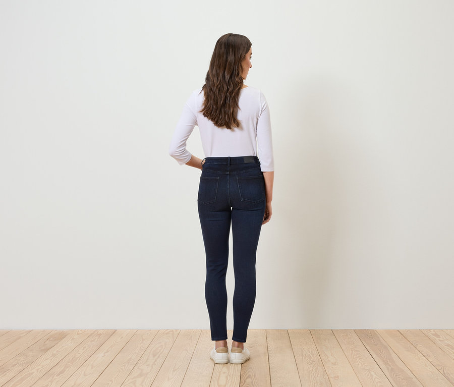 De dos, une femme aux cheveux bruns ondulés porte un jean bleu et un t-shirt blanc.