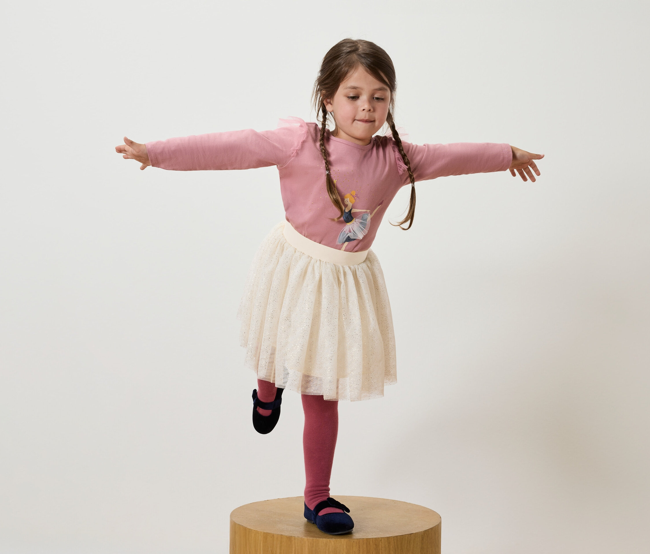 Une fille avec des tresses brunes se tient sur une jambe sur un socle en bois, portant un t-shirt rose, une jupe blanche et des chaussures bleu marine.